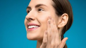 Close-up left-side portrait of a smiling woman touching her cheek against a blue background.