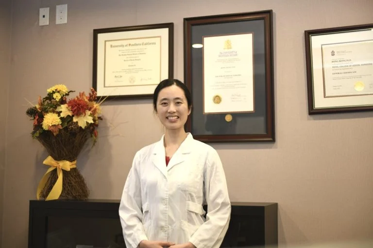 Dr. Cindy Li standing and smiling in a white coat in front of framed certificates at Excel Dental.