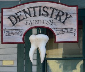 Wooden dentistry sign reading "Dentistry Painless" with a large hanging tooth below.