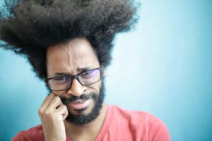 Man wearing glasses presses his cheek with a pained expression against a blue background