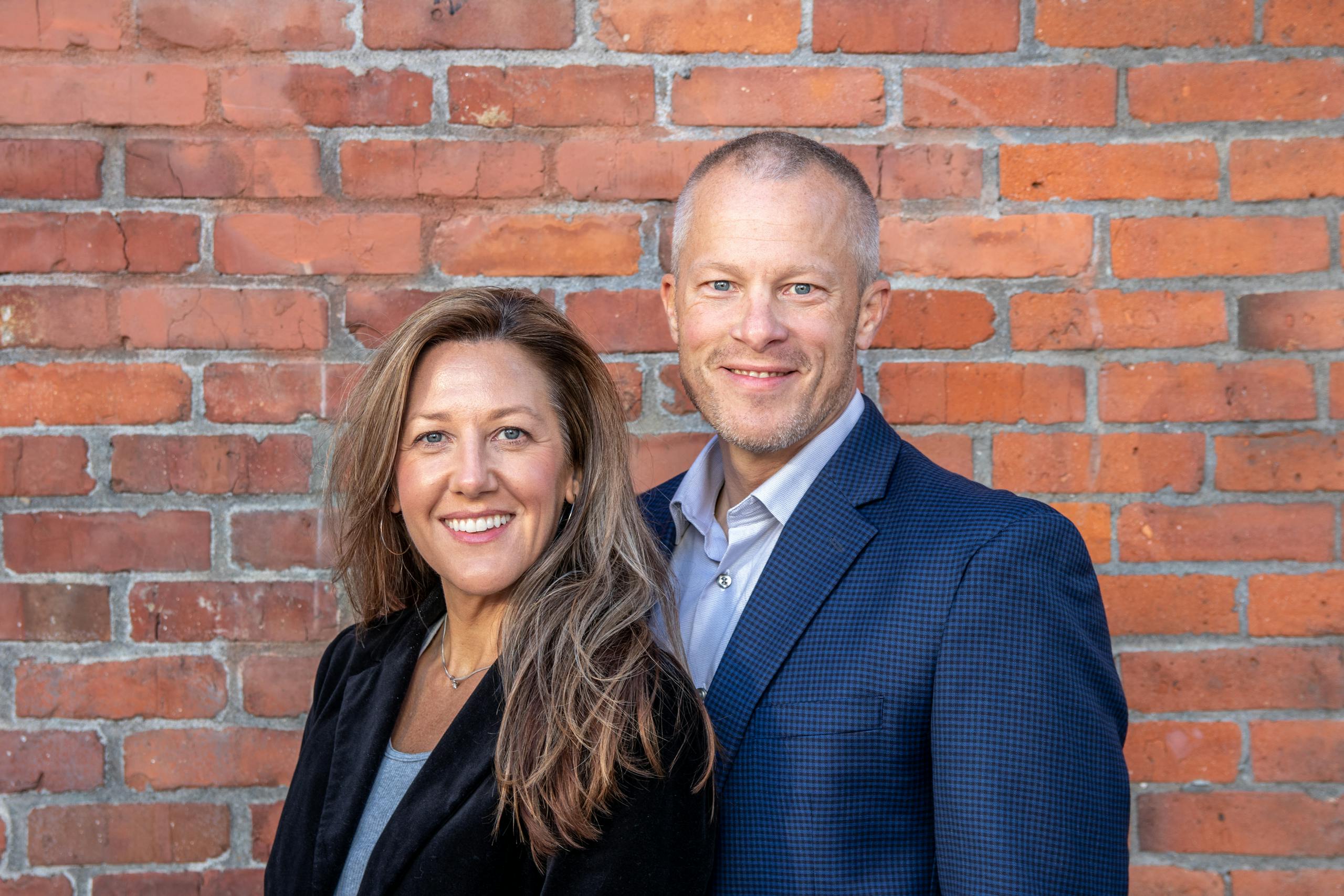 Smiling middle-aged man and woman in business attire standing in front of a brick wall.