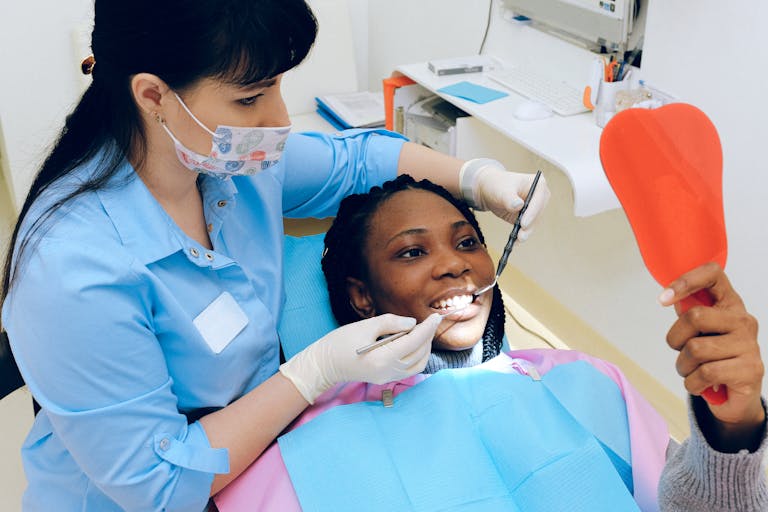 Dentist examining a patient’s teeth during a dental appointment while the patient holds a hand mirror.
