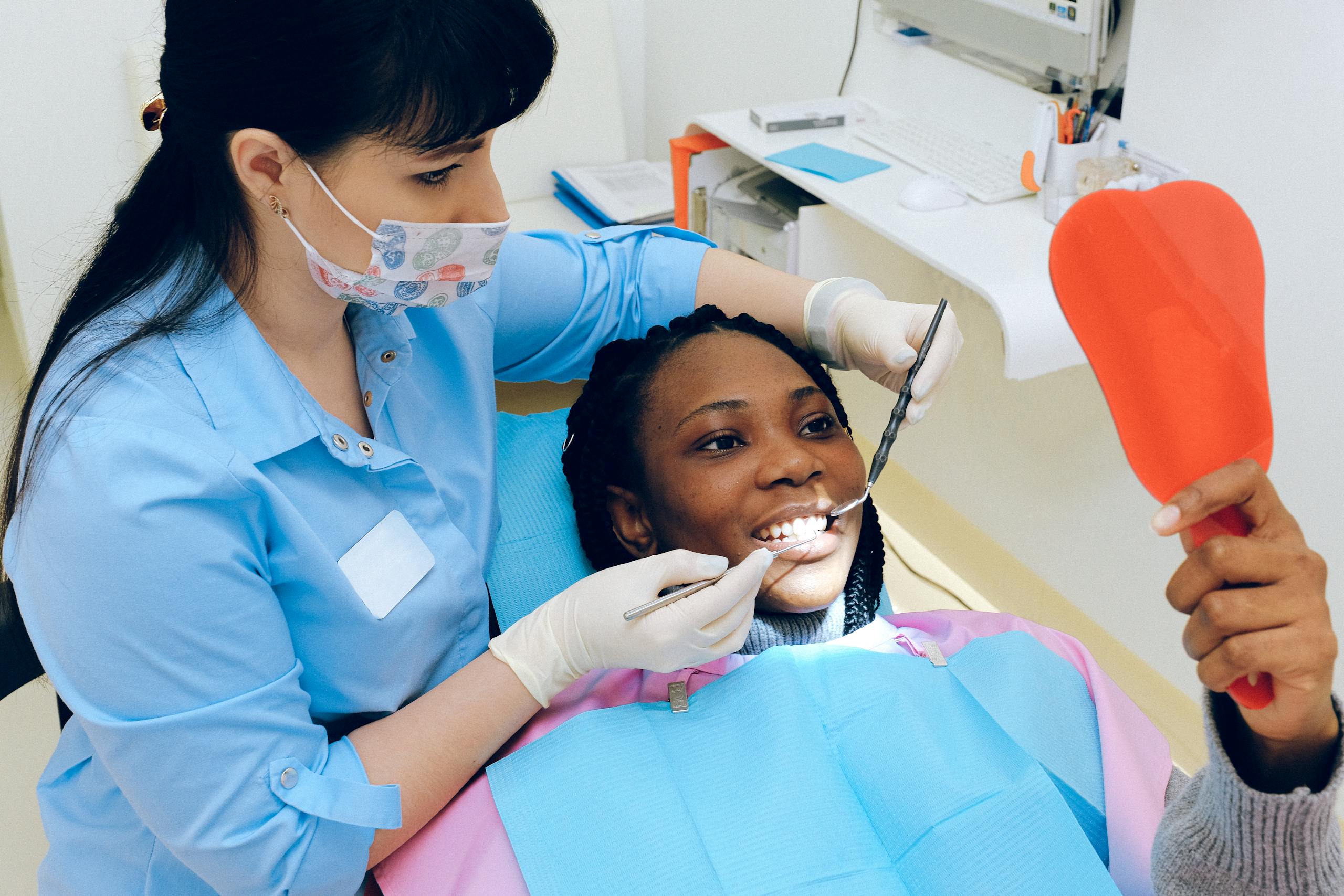 Dentist examining a patient’s teeth during a dental appointment while the patient holds a hand mirror.