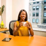 Smiling woman holding a coffee mug at an office desk near a window.