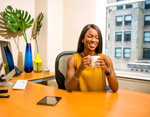 Smiling woman holding a coffee mug at an office desk near a window.