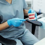 Person in scrubs and gloves pointing to a teeth model during a dental care explanation.