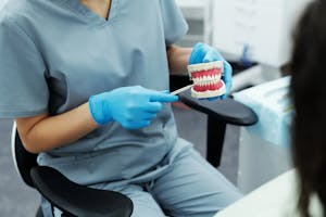 Person in scrubs and gloves pointing to a teeth model during a dental care explanation.