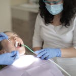 Child receiving dental care in a clinic chair while a dental professional uses instruments and an adult companion looks on.