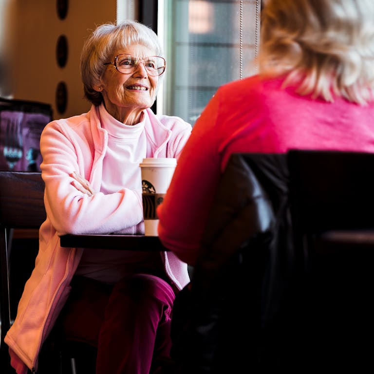 Senior woman smiling while talking with another woman at a cafe table.