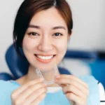 Smiling young woman in a dental chair holding a clear aligner tray.
