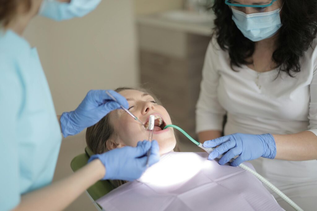 Child receiving dental care in a clinic chair while a dental professional uses instruments and an adult companion looks on.