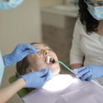 Child receiving dental care in a clinic chair while a dental professional uses instruments and an adult companion looks on.